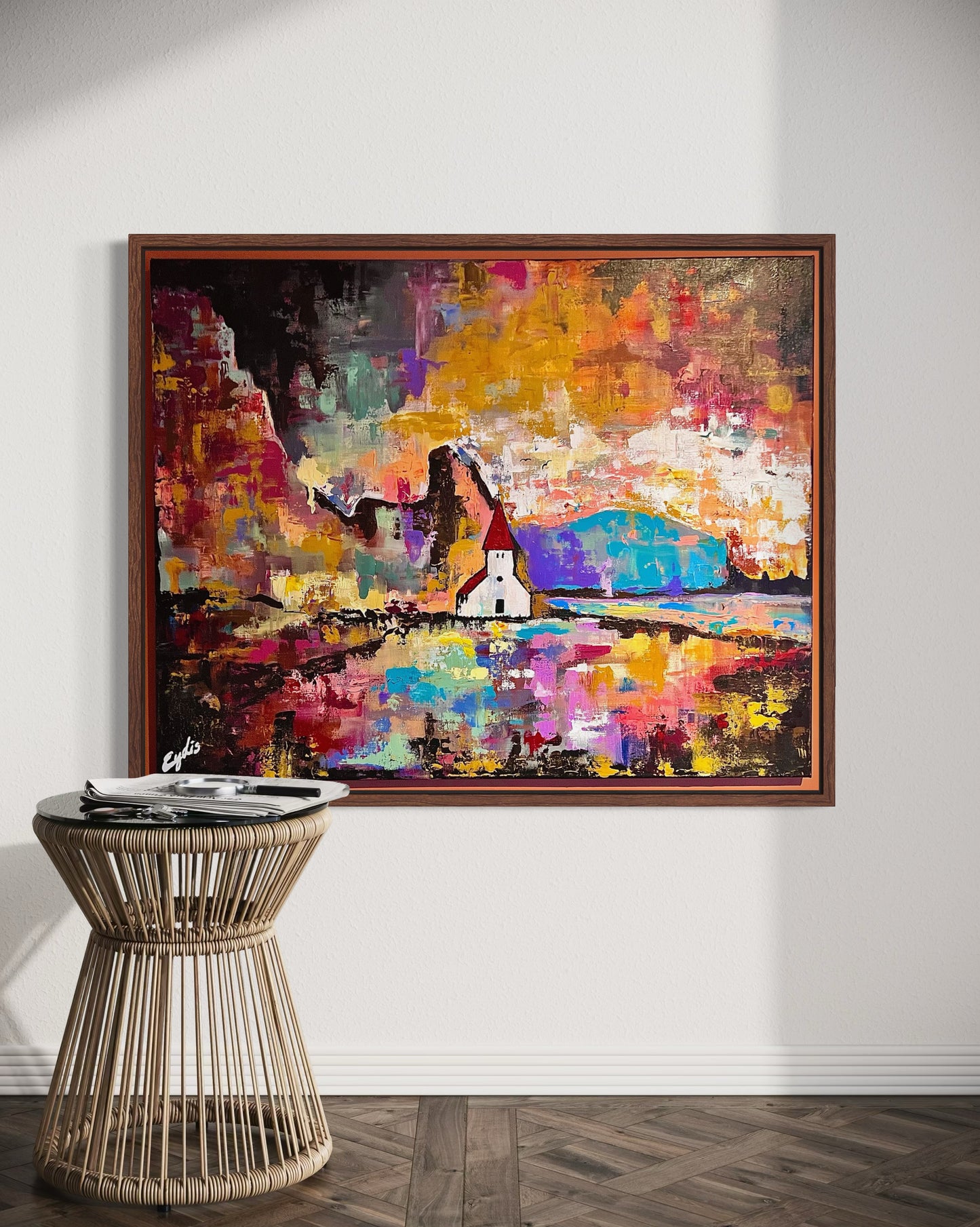 Colorful abstract Icelandic painting on a wall above a wicker side table.